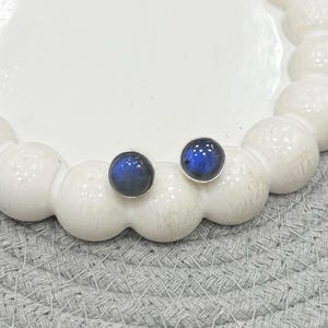 Puede incluir: Dos pendientes de botón redondos con un aspecto azul oscuro e iridiscente. Los pendientes están engarzados en metal color plata y descansan sobre un plato de cerámica blanco en forma de nube. El fondo es una superficie gris texturizada.