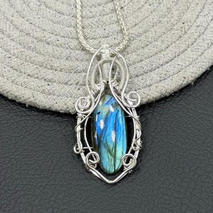 Può includere: Una collana con pendente in argento con una pietra di labradorite a forma di goccia. La pietra mostra iridescenza blu e verde, incastonata in un'incastonatura a filo dettagliata. Il pendente è appeso a una delicata catena d'argento, design di gioielli fatti a mano.
