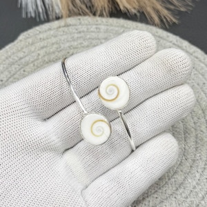 Può includere: Bracciale argentato con due ciondoli circolari bianchi con un motivo a spirale al centro. Il bracciale ha un design semplice e aperto. I ciondoli sono presentati su una superficie bianca. Ogni ciondolo misura circa 2 cm di diametro.