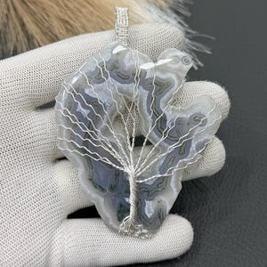 May include: A leaf-shaped pendant made from a natural stone with grey, white, and green patterns. A silver wire tree design is wrapped around the stone. The pendant is held by a gloved hand.