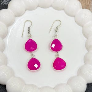 Peut inclure: Une paire de boucles d'oreilles pendantes avec deux pierres précieuses roses vives en forme de goutte d'eau. Les pierres sont serties dans du métal argenté et suspendues à un crochet simple. Les boucles sont présentées sur une assiette blanche à bord festonné.