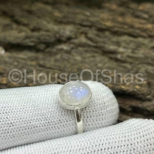 以下が含まれることがあります： 大きな丸い虹色のムーンストーンをあしらったシルバーリング。ムーンストーンは、乳白色のベースに青と白の輝きがあります。リングはシンプルで細いシルバーバンドにセットされています。「HouseOfShas」の文字が見えます。
