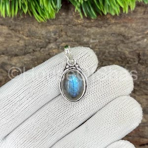 May include: Oval-shaped labradorite pendant with a silver-colored setting. The gemstone displays blue and gray iridescent colors. The pendant is suspended from a small loop at the top. The background includes green foliage and a wooden surface.