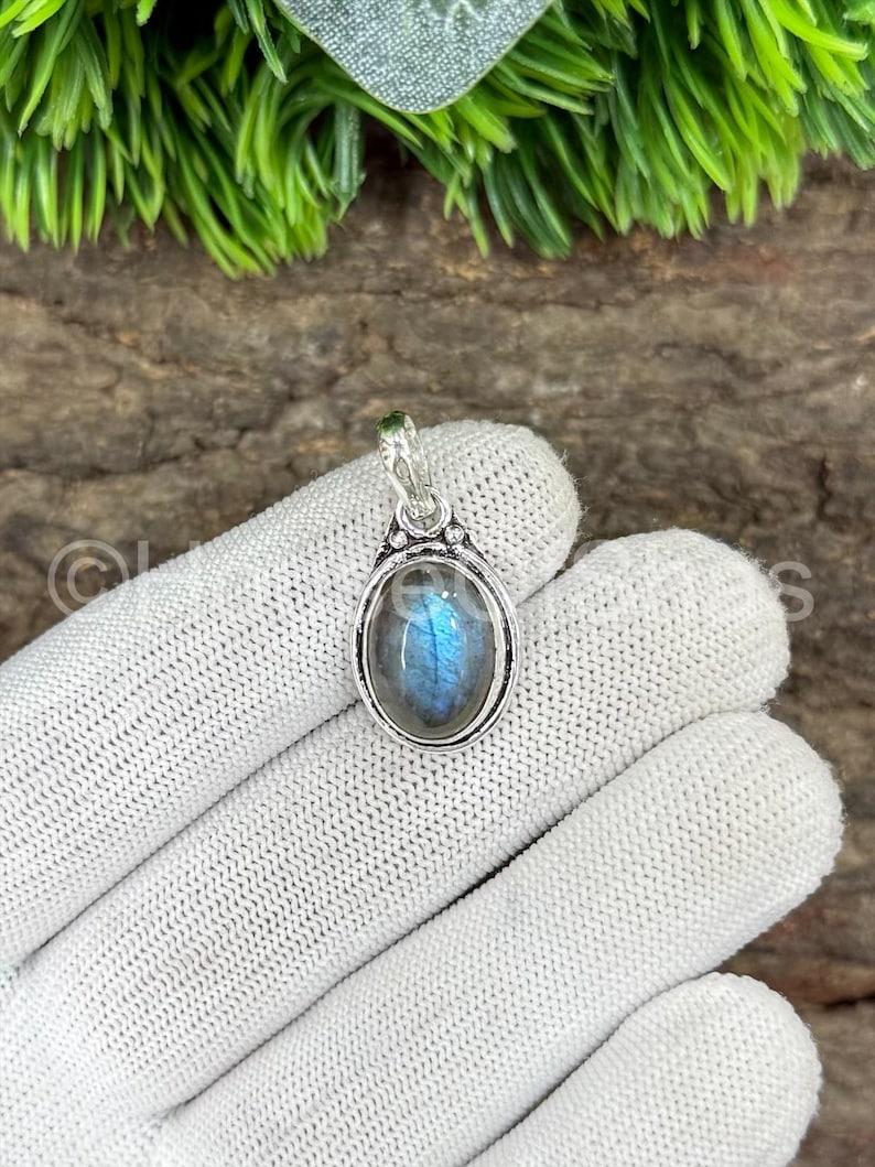 May include: Oval-shaped labradorite pendant with a silver-colored setting. The gemstone displays blue and gray iridescent colors. The pendant is suspended from a small loop at the top. The background includes green foliage and a wooden surface.