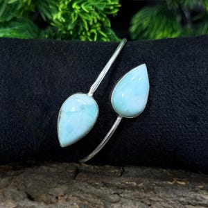 Puede incluir: Un brazalete plateado con dos piedras de larimar azul pálido en forma de lágrima. El brazalete tiene un diseño abierto, con las piedras colocadas en los extremos opuestos de la banda. Las piedras tienen una superficie pulida y el brazalete se muestra sobre una superficie negra. Mide unos 15 cm.