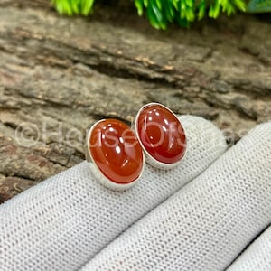May include: A pair of oval stud earrings. Each earring has a polished, reddish-orange gemstone set in silver-coloured metal. The earrings are presented on a white, textured surface.