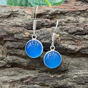 Peut inclure: Paire de boucles d'oreilles pendantes argentées. Chaque boucle présente une pierre ronde bleu vif sertie dans un cadre argenté. Les boucles sont suspendues à un crochet simple avec un design en spirale. Fond texturé, marron.