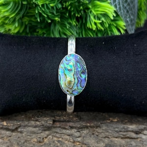 May include: Silver bracelet with an oval abalone shell centrepiece. The shell has iridescent blues, greens, and purples. The bracelet is set against a black background, with greenery behind it. Abalone shell jewellery.