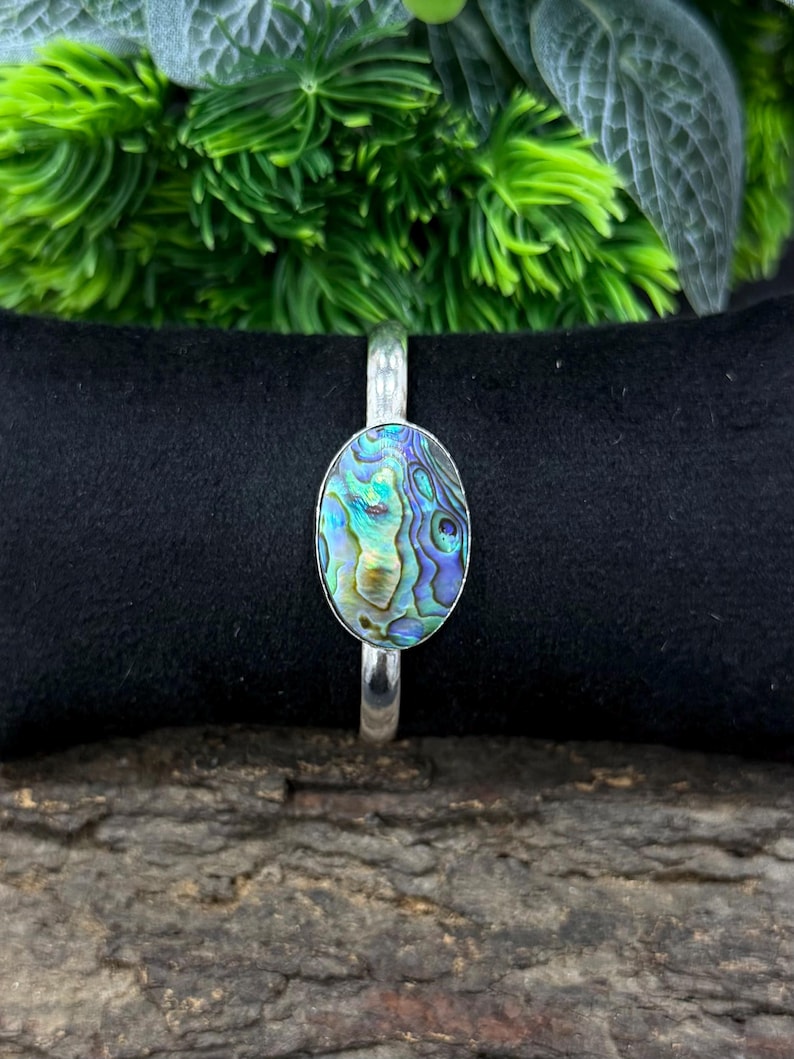 Pode incluir: Pulseira prateada com um centro oval de concha de abalone. A concha exibe iridesc&ecirc;ncias azuis, verdes e roxas. A pulseira &eacute; colocada sobre um fundo preto, com vegeta&ccedil;&atilde;o ao fundo. Joias de concha de abalone.