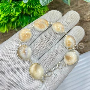 Op de afbeelding: Een zilveren armband met ovale en ronde, cr&egrave;mekleurige cabochonstenen. De stenen zijn gezet in een zilveren ketting met een sluiting. De armband wordt gepresenteerd op een wit oppervlak.