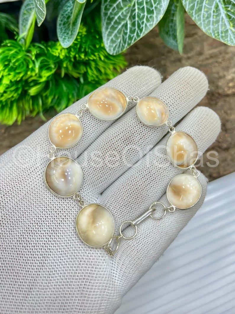 Op de afbeelding: Een zilveren armband met ovale en ronde, cr&egrave;mekleurige cabochonstenen. De stenen zijn gezet in een zilveren ketting met een sluiting. De armband wordt gepresenteerd op een wit oppervlak.