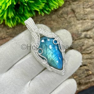 May include: A handcrafted silver pendant featuring a blue labradorite gemstone. The gemstone is wire-wrapped in a detailed, ornate design. The pendant has a teardrop shape and is displayed against a neutral background.