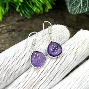 May include: A pair of teardrop-shaped amethyst earrings. Each earring features a polished, deep purple gemstone set in a silver bezel. The earrings have a simple hook design with a coiled accent above the gemstone. The background is a white glove.