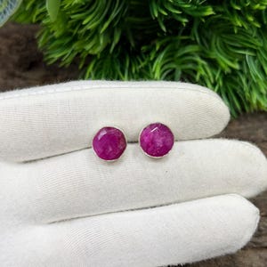 Op de afbeelding: Een paar ronde, magenta edelsteen oorknopjes. De oorbellen zijn gezet in zilver en worden gepresenteerd op een wit oppervlak. Groen gebladerte is zichtbaar op de achtergrond.