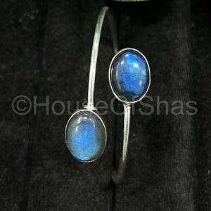 May include: A silver bracelet with two oval labradorite stones, displaying iridescent blue hues. The stones are set in a bezel setting and connected by a slender, open cuff. The bracelet is shown against a dark backdrop.
