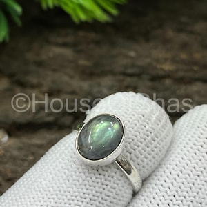 以下が含まれることがあります： 楕円形のラブラドライト宝石をあしらったシルバーリング。虹色の石は緑と青の色合いを示しています。リングは白い表面にセットされています。