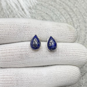 Puede incluir: Un par de pendientes de botón en forma de lágrima con piedras de lapislázuli azul intenso y sutiles inclusiones de pirita dorada, cada una engastada en un marco de metal plateado pulido, con una superficie lisa, redondeada y clásica.