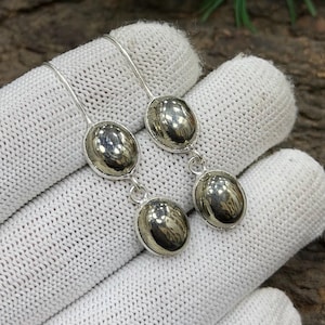 Pode incluir: Brincos pendurados em prata com três pedras ovais de pirita metálica. Os brincos são apresentados em um tecido branco texturizado, com um fundo de madeira e folhas verdes.