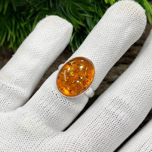 Peut inclure: Bague en argent ornée d'une pierre d'ambre ovale avec des inclusions dorées. La bague est présentée sur une surface blanche, avec un fond de feuillage vert. La pierre d'ambre mesure environ 2,5 cm de long.