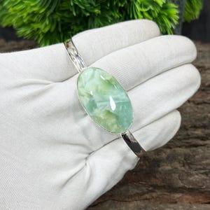 May include: A silver bracelet featuring an oval, light green gemstone. The gemstone has a marbled appearance with shades of white and green. The bracelet is held by a gloved hand against a backdrop of greenery and wood.