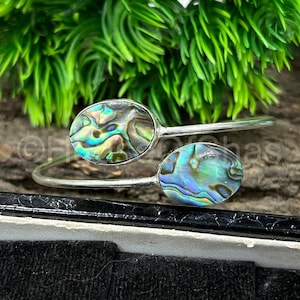 Puede incluir: Una pulsera de puño plateada con dos incrustaciones ovaladas de concha de abulón. Las conchas iridiscentes muestran una mezcla de azules, verdes y morados. La pulsera está colocada sobre un fondo de follaje verde y una superficie oscura.