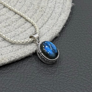 Peut inclure: Un collier pendentif en argent avec une pierre précieuse labradorite de forme ovale aux reflets bleutés. La pierre est sertie dans une monture en argent décorative avec des motifs de volutes complexes sur le côté, sur une chaîne tressée.