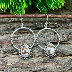 May include: A pair of silver hoop earrings with a teardrop-shaped stone pendant. The stone has a white and brown pattern, and the pendant is framed by a beaded silver border. The earrings are displayed against a textured wooden background.