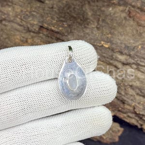 May include: A silver pendant with a teardrop shape, featuring a small, oval, light-colored stone. The pendant is held against a white, textured surface, with a wooden background visible. The pendant has a loop at the top for a chain.