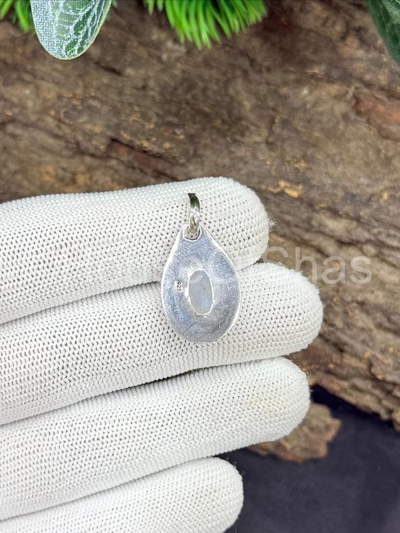 May include: A silver pendant with a teardrop shape, featuring a small, oval, light-colored stone. The pendant is held against a white, textured surface, with a wooden background visible. The pendant has a loop at the top for a chain.