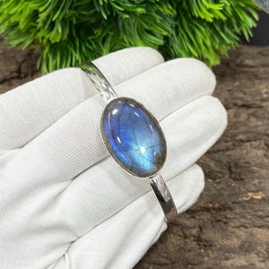 May include: A silver bracelet featuring an oval-shaped, iridescent blue gemstone. The gemstone displays a play of colors, with flashes of blue and green. The bracelet is set against a white background, highlighting the jewelry's design.