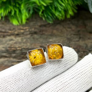 May include: A pair of square amber stud earrings. Each earring has a polished, golden-yellow amber stone set in a silver-coloured metal frame. The amber has visible inclusions. The earrings are displayed on a white surface.