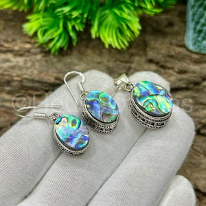 May include: Abalone shell jewelry set featuring a pair of earrings and a pendant. The oval-shaped abalone shells display iridescent blues, greens, and purples, set in ornate silver-toned metal. The earrings have a hook closure.