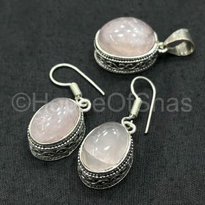May include: A jewelry set featuring a pendant and a pair of earrings. Each piece has a light pink, oval-shaped stone set in a silver-colored metal frame with an ornate design. The pendant has a loop for a chain.