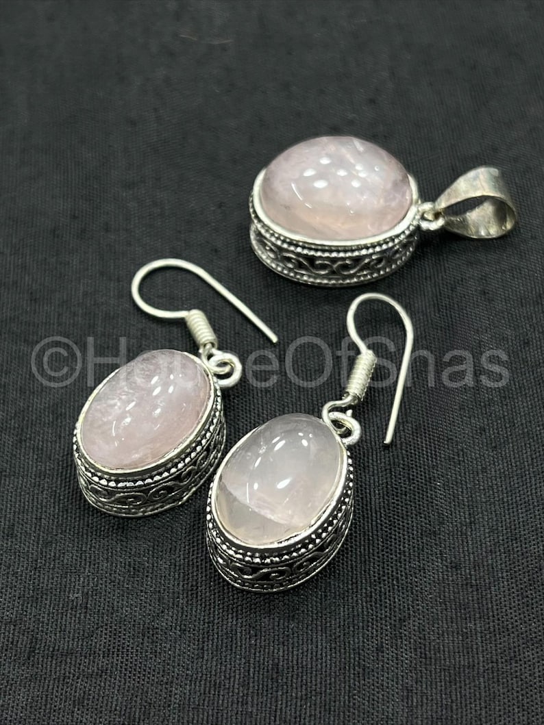 May include: A jewelry set featuring a pendant and a pair of earrings. Each piece has a light pink, oval-shaped stone set in a silver-colored metal frame with an ornate design. The pendant has a loop for a chain.