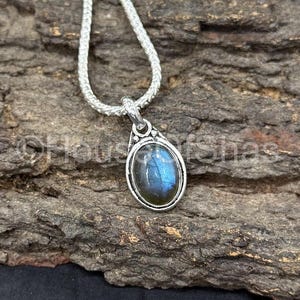 May include: A silver pendant necklace featuring an oval labradorite gemstone with blue and green iridescence. The pendant is set in a silver bezel and hangs from a silver chain. The necklace is displayed on a textured, brown surface.