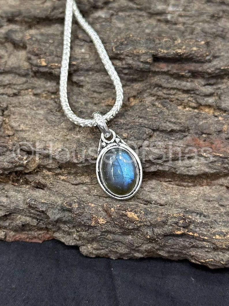 May include: A silver pendant necklace featuring an oval labradorite gemstone with blue and green iridescence. The pendant is set in a silver bezel and hangs from a silver chain. The necklace is displayed on a textured, brown surface.
