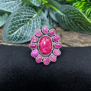 May include: A silver-toned ring featuring a large, oval-shaped, deep pink gemstone at the center, surrounded by smaller, oval-shaped pink stones. The ring has a floral-like design, with the smaller stones arranged around the central stone.