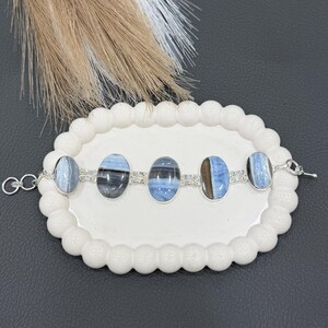 Op de afbeelding: Een zilveren armband met vijf ovale, blauw-grijze edelstenen. De armband wordt gepresenteerd op een wit bord met geschulpte rand. De edelstenen zijn verbonden door zilveren schakels. De achtergrond is donker.