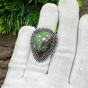 Może przedstawiać: Srebrny pierścionek z dużym, łzowym kamieniem szlachetnym. Kamień szlachetny to mieszanka zielonych i czerwonych odcieni, osadzona w szczegółowej, koralikowej srebrnej ramie. Pierścionek jest prezentowany na białej rękawicy, z zielonymi liśćmi w tle.