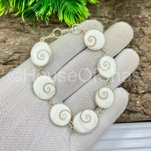 May include: A silver bracelet featuring oval and round white shell segments with a spiral design. The bracelet is connected by small silver links and is displayed on a white glove.