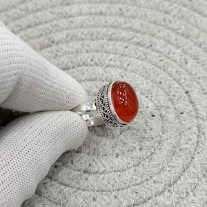 Pode incluir: Um anel de prata com uma pedra preciosa de cornalina laranja-escura, lisa e de formato oval, engastada numa moldura de metal decorativa com detalhes intrincados de volutas e missangas ao longo das bordas da cravação.