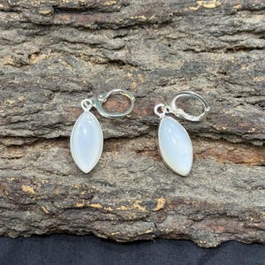May include: A pair of silver leverback earrings, each with a pear-shaped, iridescent, white gemstone. The earrings are presented on a textured, brown, bark-like surface.