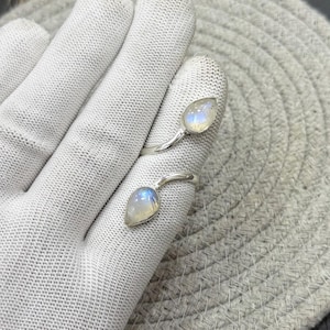 Puede incluir: Dos anillos de plata, cada uno con una piedra lunar en forma de pera. Las piedras lunares muestran un juego de colores brillante e iridiscente. Los anillos se muestran sobre un guante blanco sobre un fondo gris.