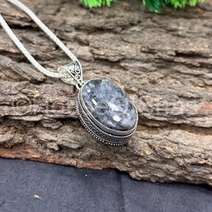 Pode incluir: Um colar com pingente de prata com uma pedra oval cinza e branca. A pedra é definida em uma moldura decorativa de prata com uma borda de contas. A corrente do colar é fina e prateada.