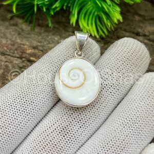 May include: A round, white pendant with a silver bail. The pendant features a spiral design in the center, with a gold-colored accent. The pendant is set in a silver bezel. The background is blurred, with green foliage.