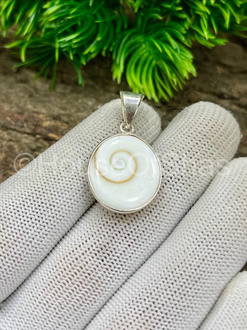 May include: A round, white pendant with a silver bail. The pendant features a spiral design in the center, with a gold-colored accent. The pendant is set in a silver bezel. The background is blurred, with green foliage.