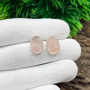 Può includere: Un paio di orecchini a lobo in quarzo rosa di forma ovale. Gli orecchini hanno una montatura color argento e una tonalità rosa tenue. Sono esposti su una superficie bianca a maglia, con un fondo di fogliame verde.