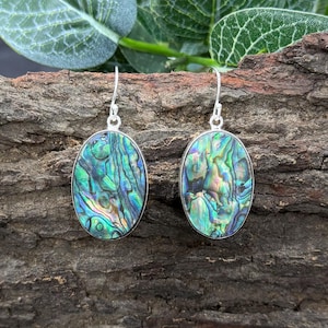 May include: A pair of oval-shaped earrings with iridescent abalone shells set in silver. The shells display a mix of green, blue, and purple hues. The earrings hang from silver hooks, showcasing a natural, organic design.