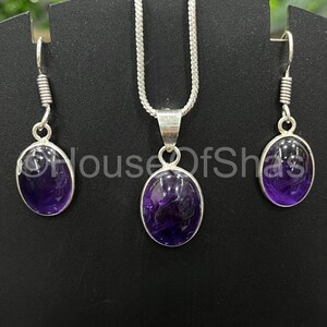 May include: A jewelry set featuring a silver necklace with a pendant and matching earrings. Each piece showcases a polished, oval-shaped, deep purple gemstone. The earrings have a hook design, and the necklace has a delicate chain.