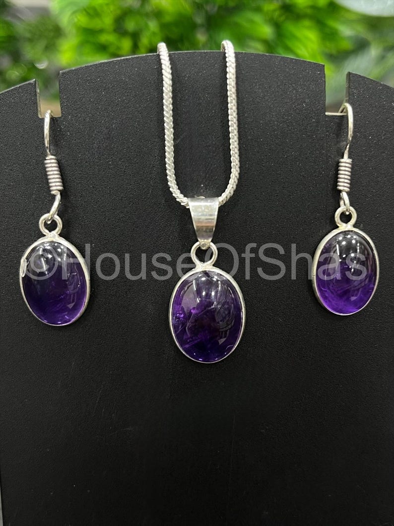 May include: A jewelry set featuring a silver necklace with a pendant and matching earrings. Each piece showcases a polished, oval-shaped, deep purple gemstone. The earrings have a hook design, and the necklace has a delicate chain.